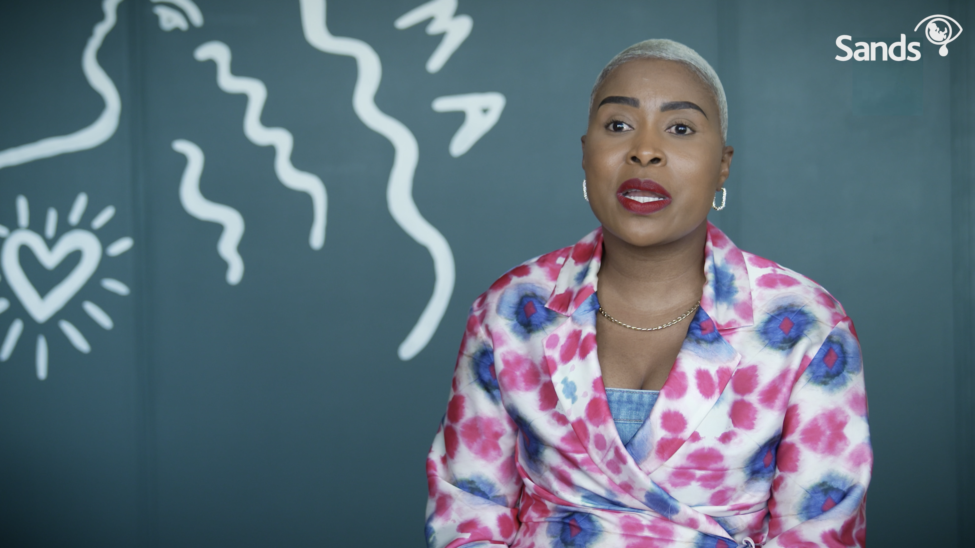 A woman in a pink and white dress is sat in front of a chalkboard background, speaking to the camera. 