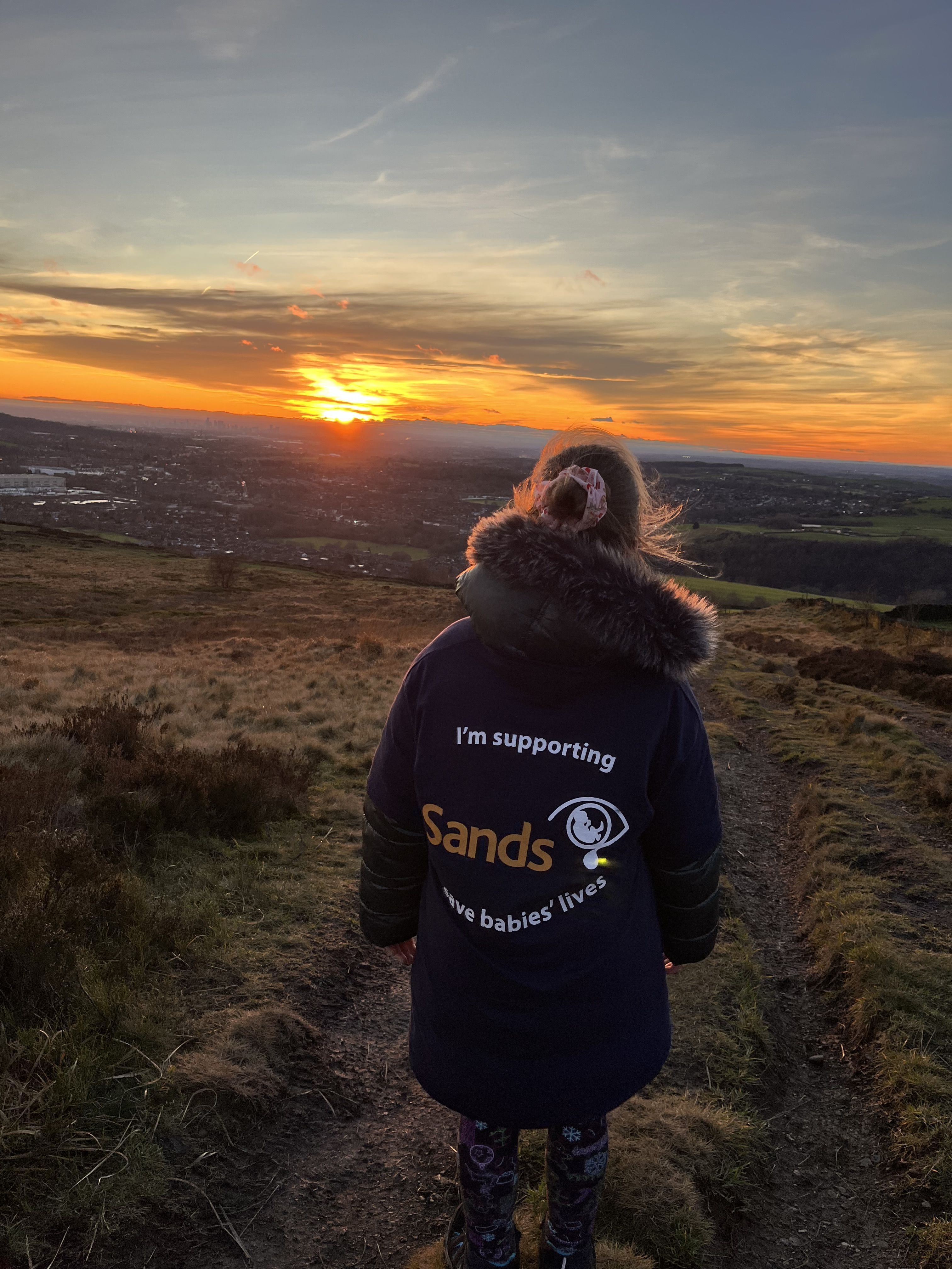 A young Sands Supporter hiking in the countryside facing towards an orange sunset. She is wearing a blue Sands T-shirt.