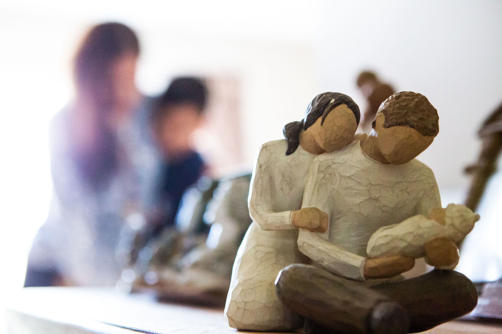 A statuette of a mother, father holding a baby. Blurred in the background is a South Asian Mother embracing a child. 