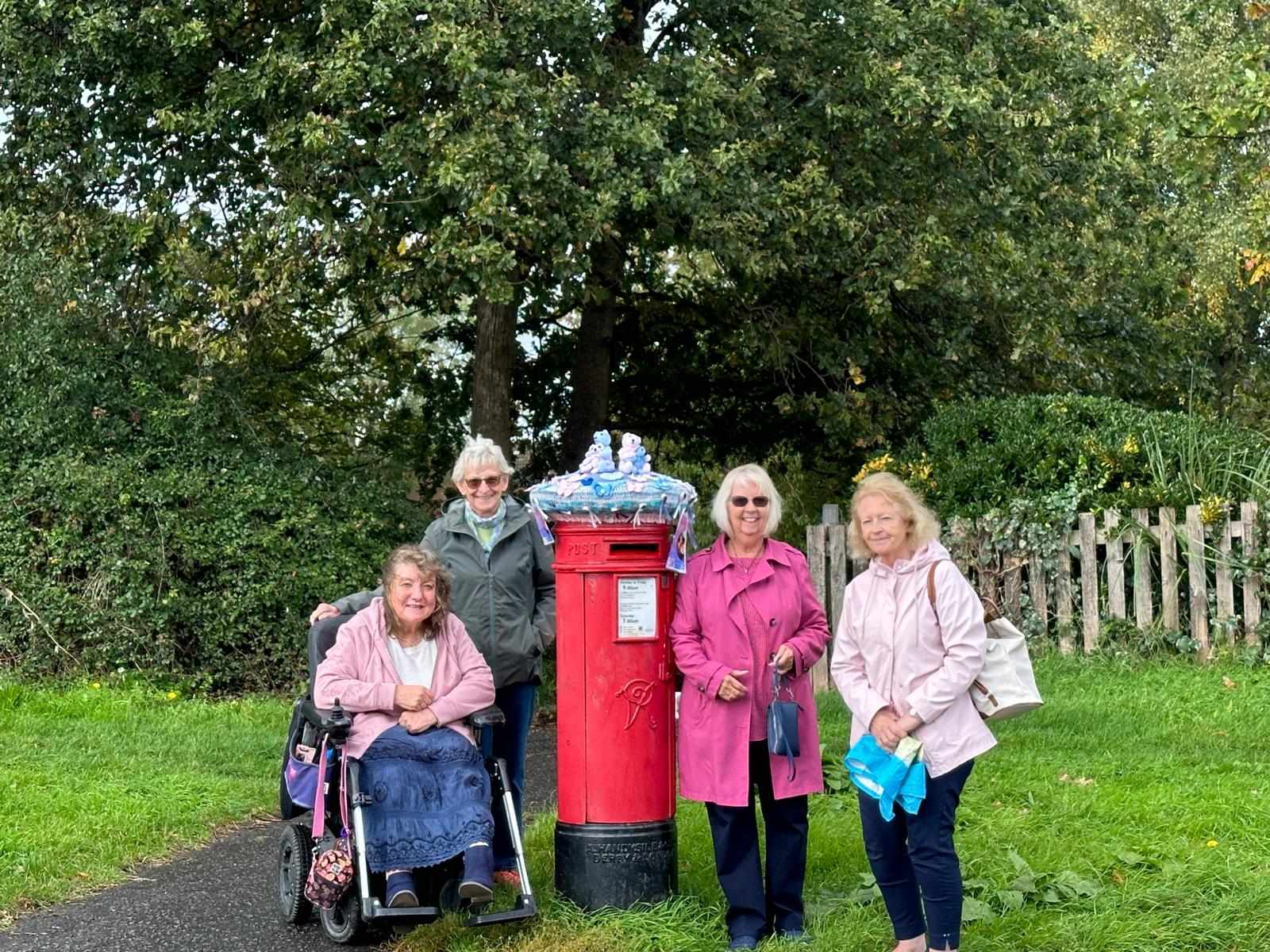 A group of women pose next to a bright red post box with a knitted topper, raising awareness for pregnancy and baby loss. 