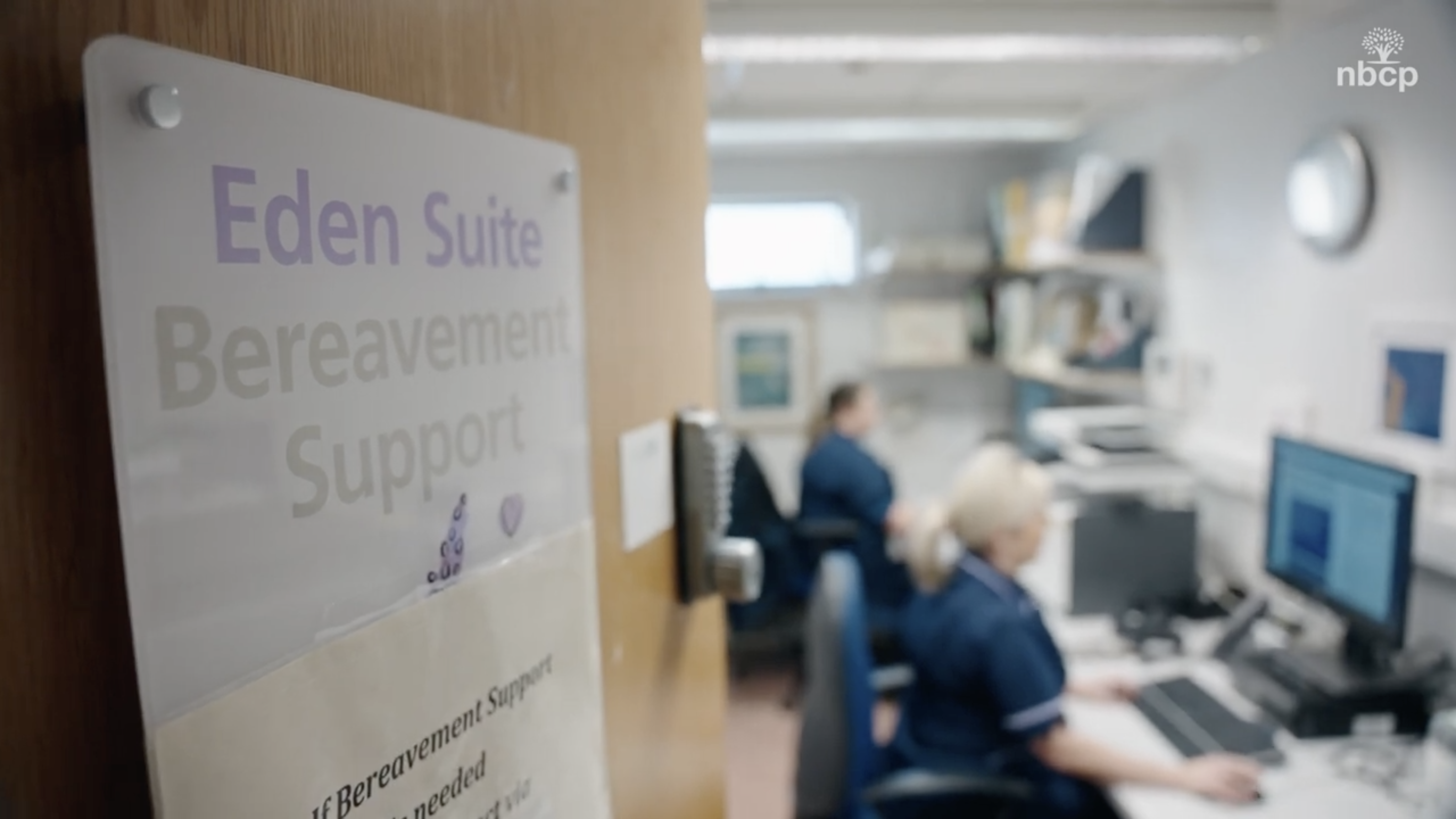 2 Bereavement Support nurses sitting in an office using computers. The door to the room is close to the camera and has a sign saying 'Eden Suite, Bereavement Support'.