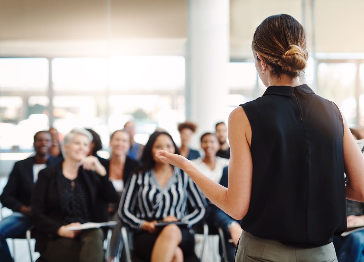 A woman is stood at the front of the room with her back to the camera, talking to a group of people. 