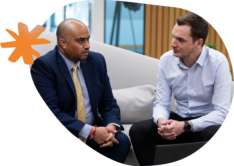 Two male work colleagues seated on a couch, discussing something intently in a relaxed office setting. 