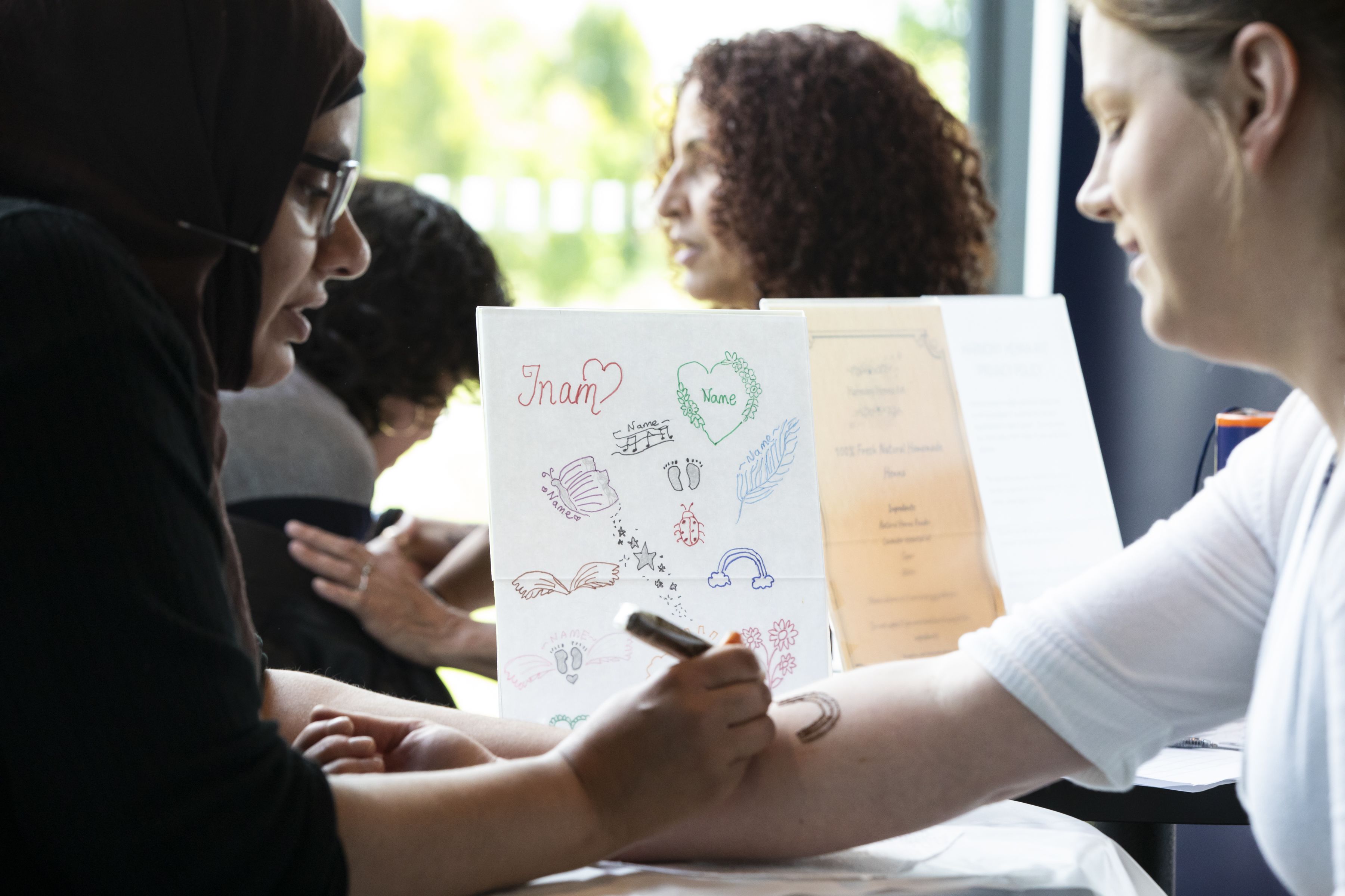 An attendee at Sands Garden Day getting a henna tattoo of a rainbow on her arm and talking with the woman applying the henna.