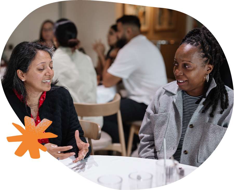 Two women are sat together at a table and engaged in conversation in a room full of people sharing their experiences. 