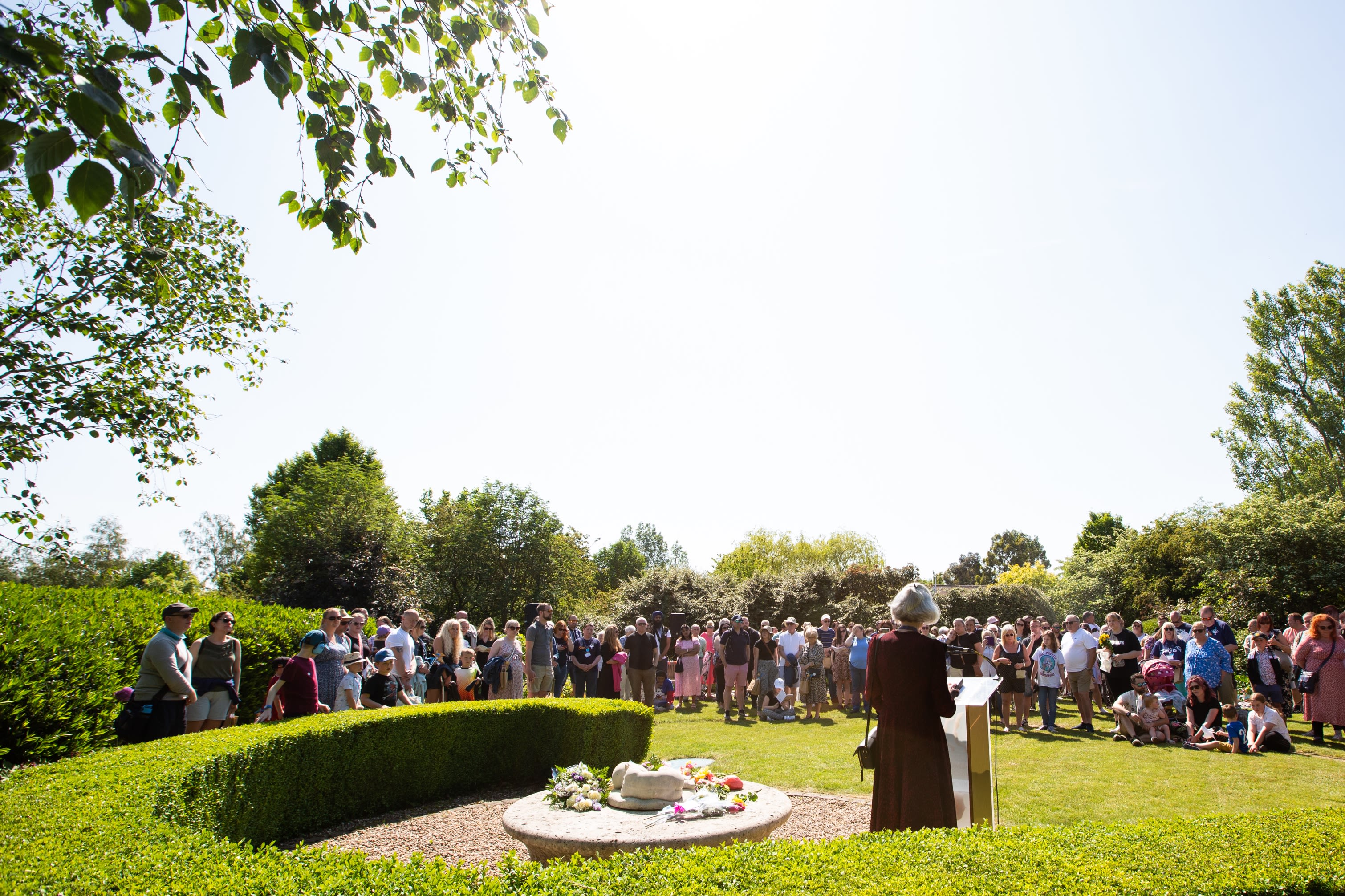 Clea Harmer speaking in front of a large group of bereaved families and supporters at the Sands Garden Day in 2023. They are standing in the Sands Garden on a bright and sunny day.