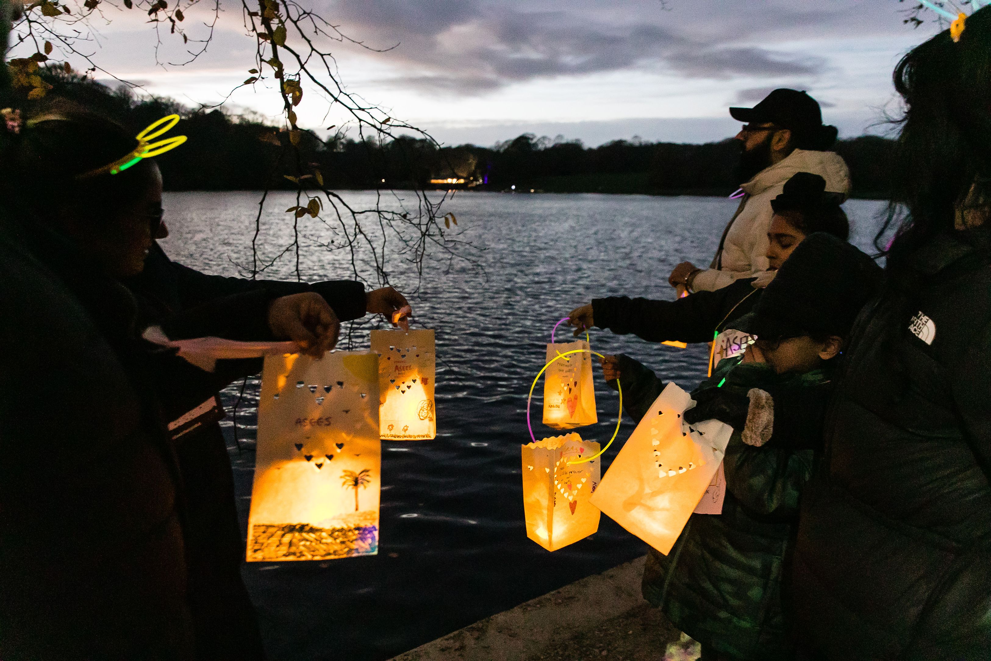 A group of people holding paper lanterns next to a lake as the sun sets in memory of their much-loved babies as part of a Sands Starlight Walk event. 
