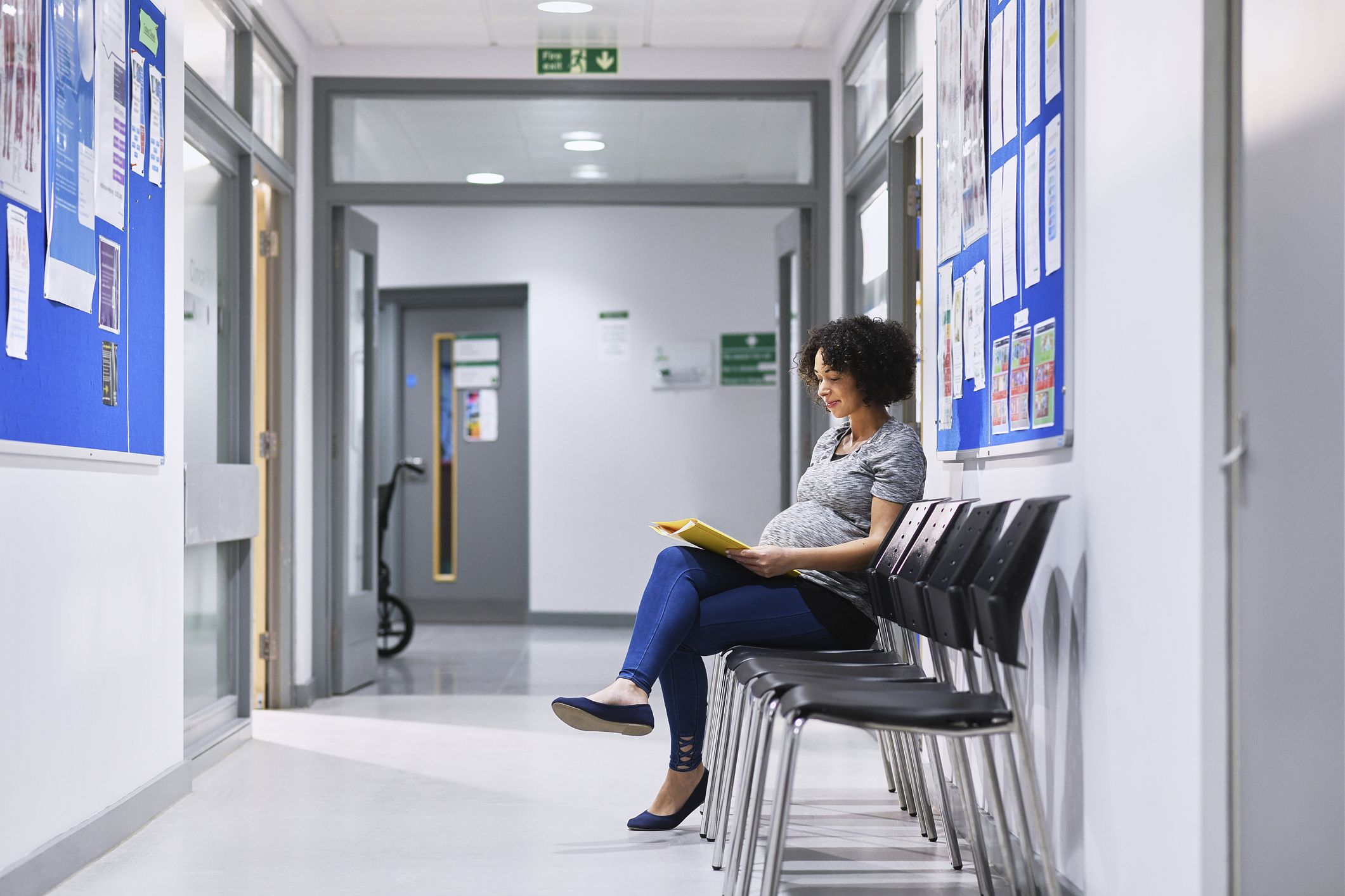A pregnant woman seated on a chair in a corridor of a health care building, dressed casually and appearing relaxed reading paperwork.