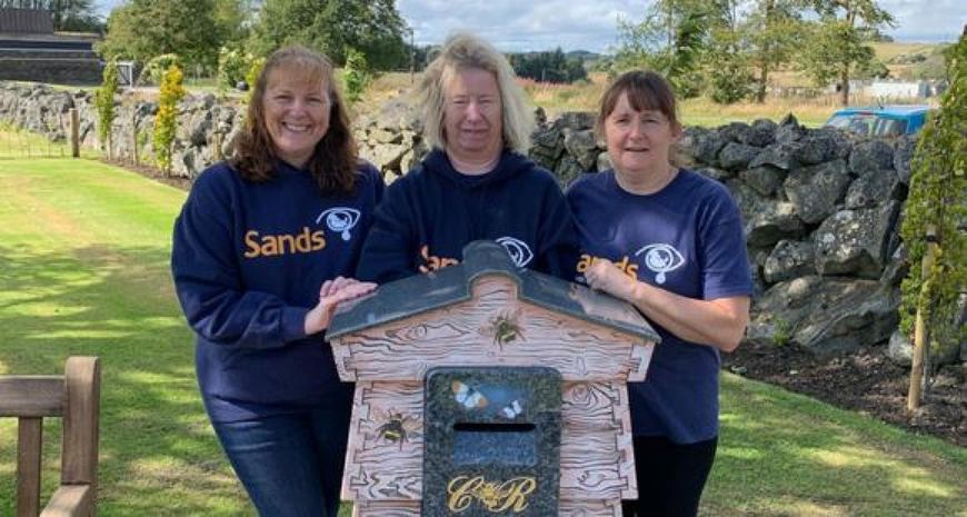 Three women stood together around a bee hive wearing Sands t-shirts. 