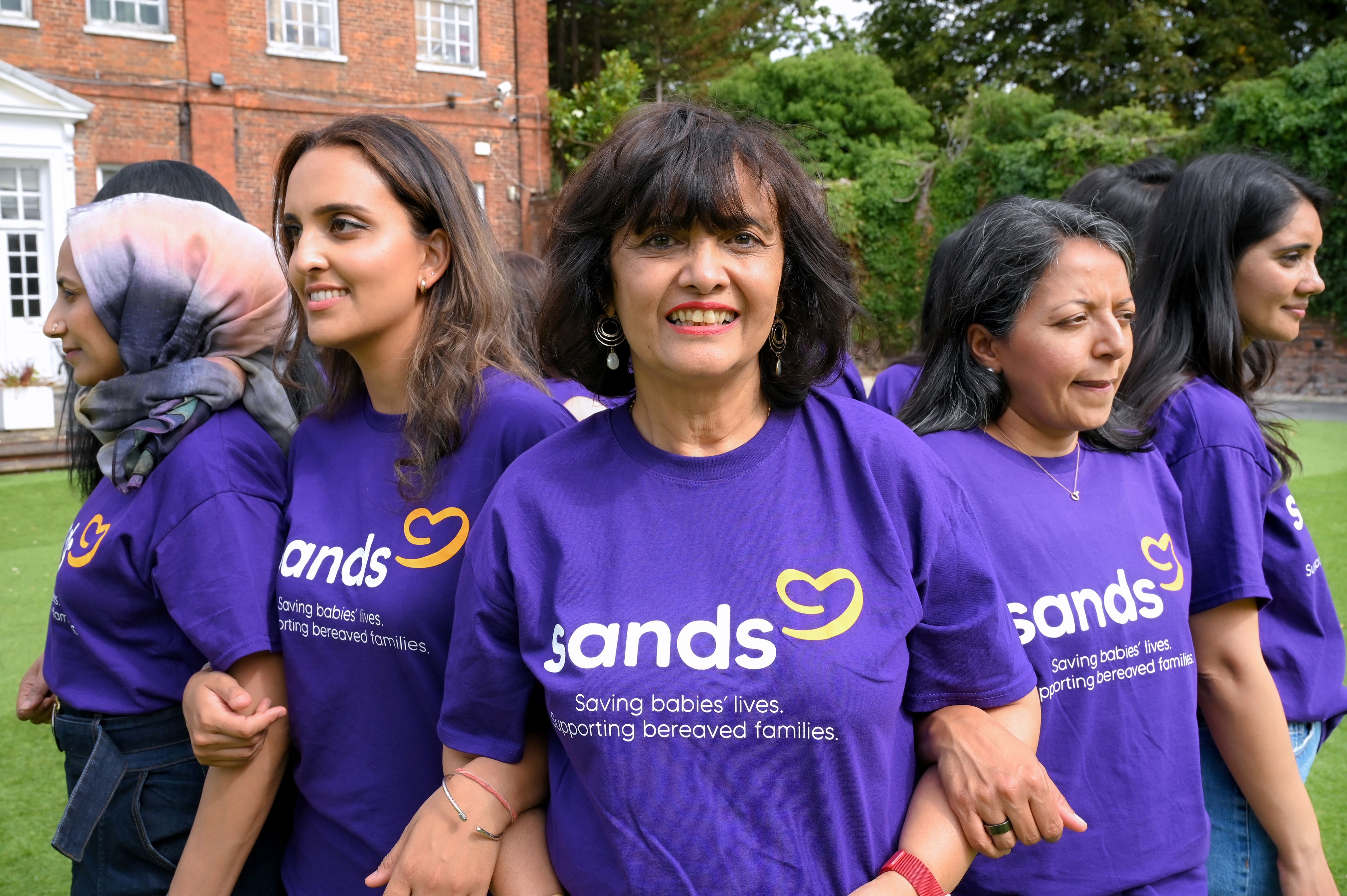 A group of South Asian women are stood together in a circle, wearing Sands t-shirts, and linking arms whilst looking forward.