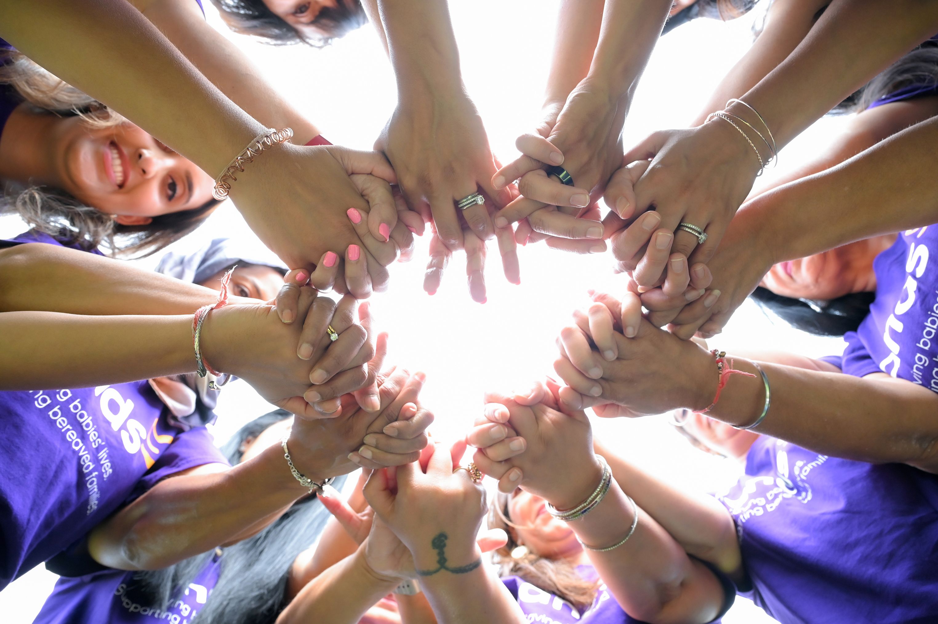 A group of South Asian women stood together in a circle, smiling and holding hands as they look down towards the camera. 
