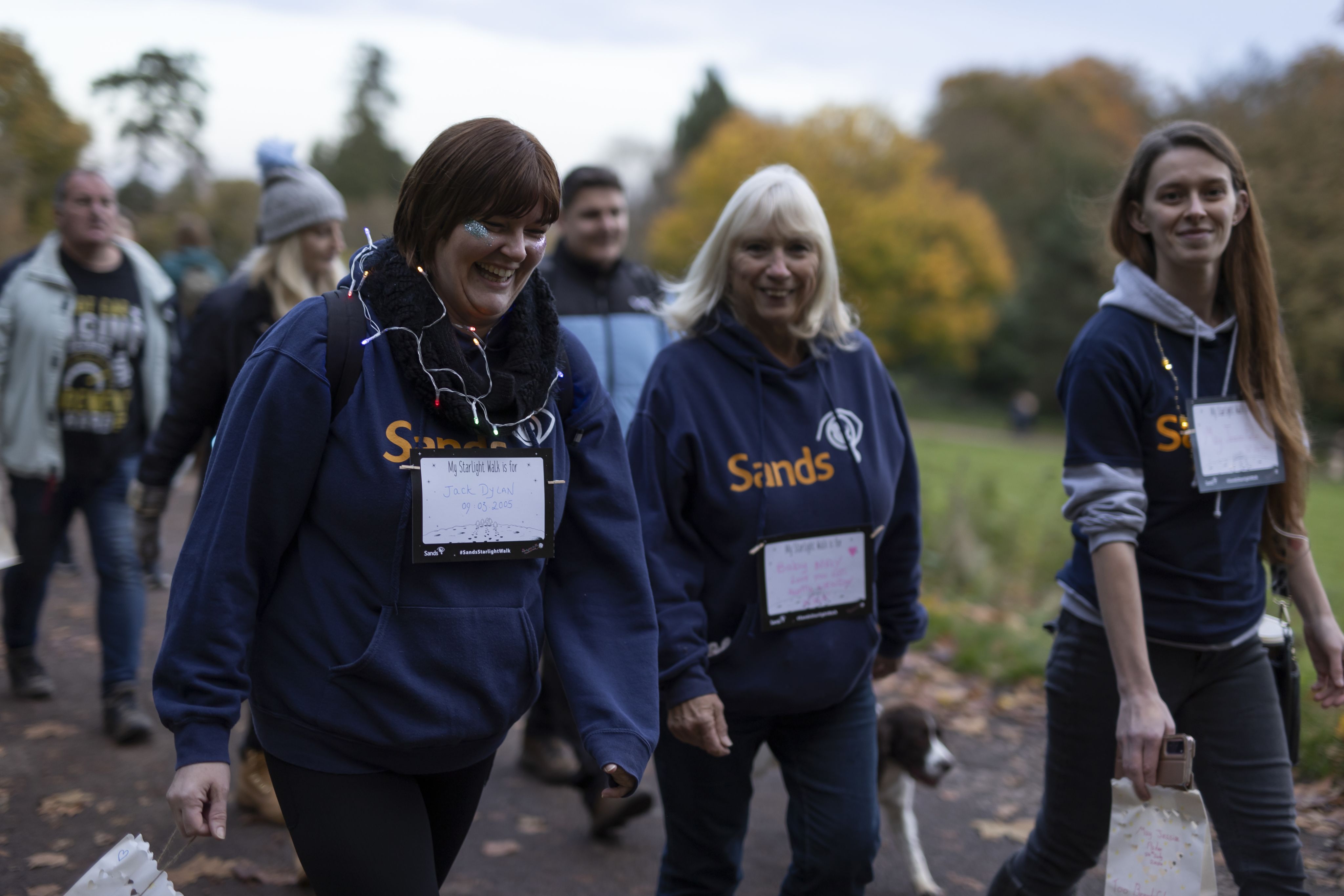 3 female Sands supporters walking and smiling in remembrance at a Starlight Walk event. They are holding paper lanterns and are wearing blue Sands hoodies.