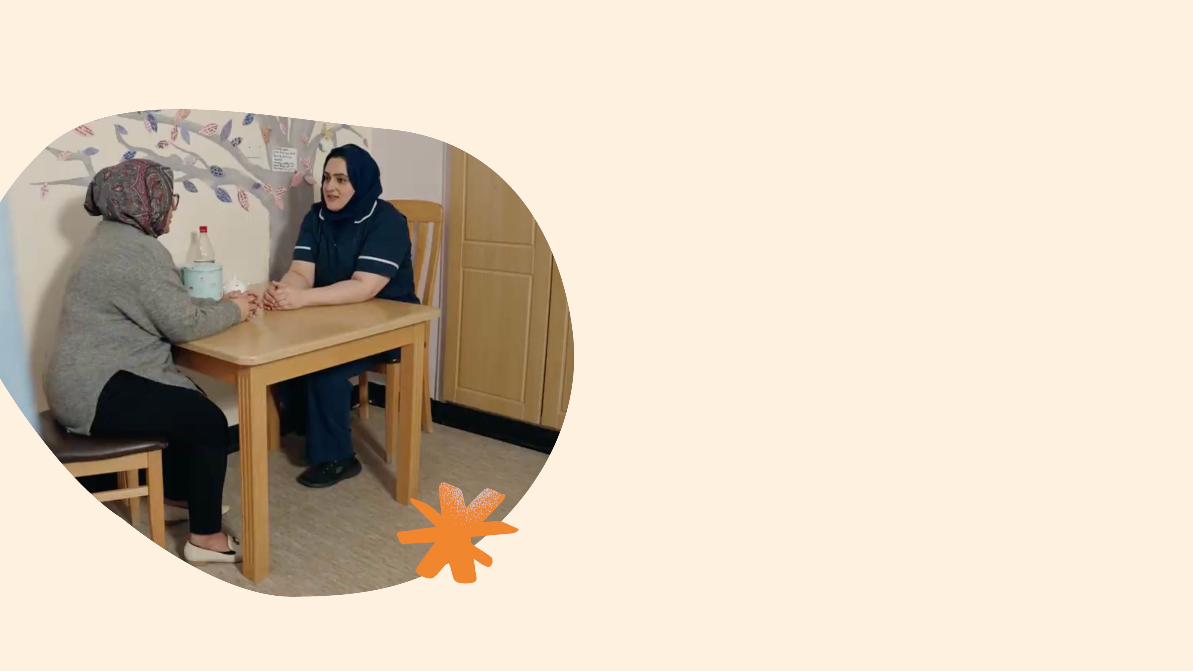 A nurse is sat opposite a parent at a table in a hospital room, engaged in conversation. 