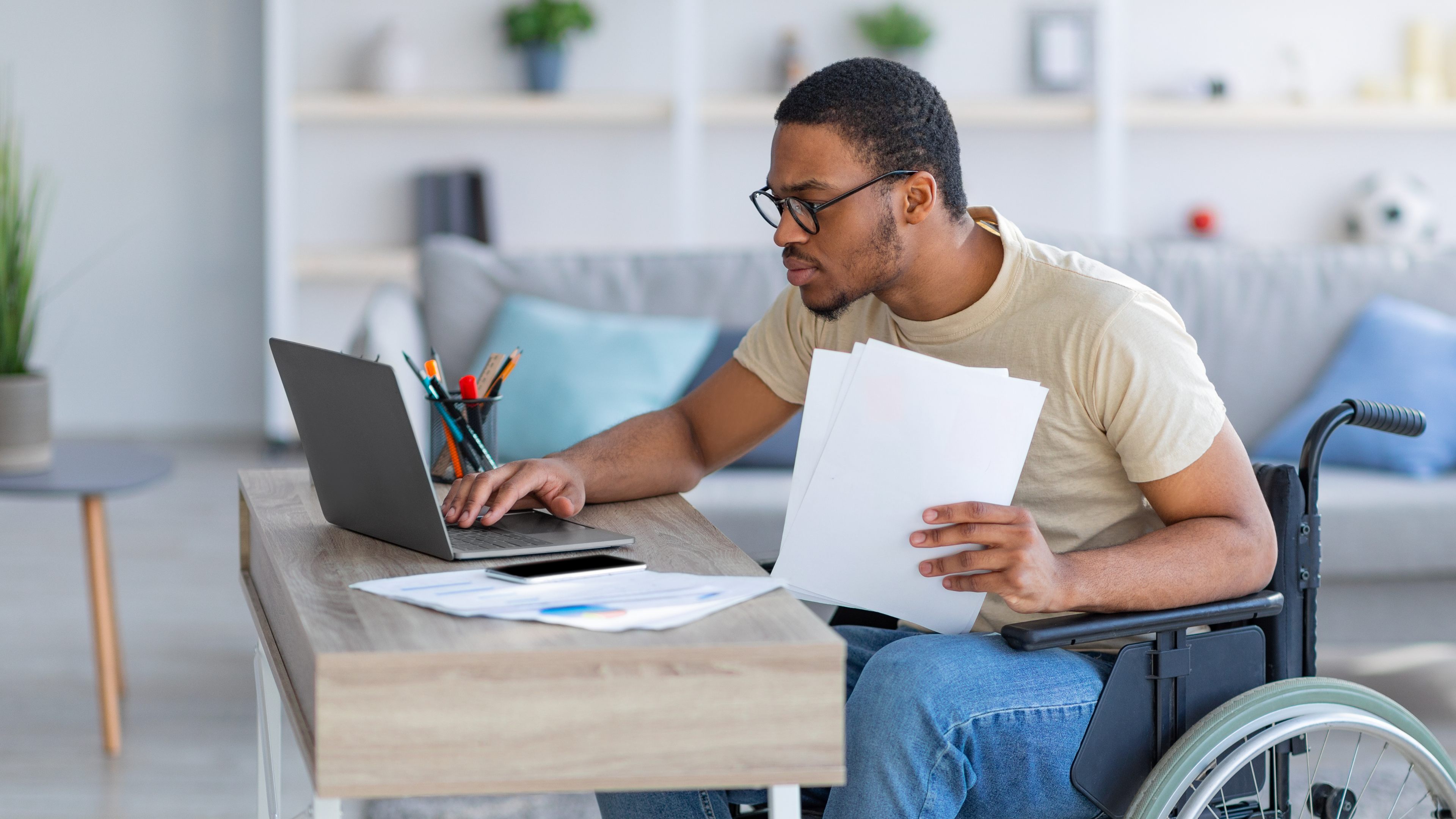 Man in wheelchair sits at table on laptop, holding papers.