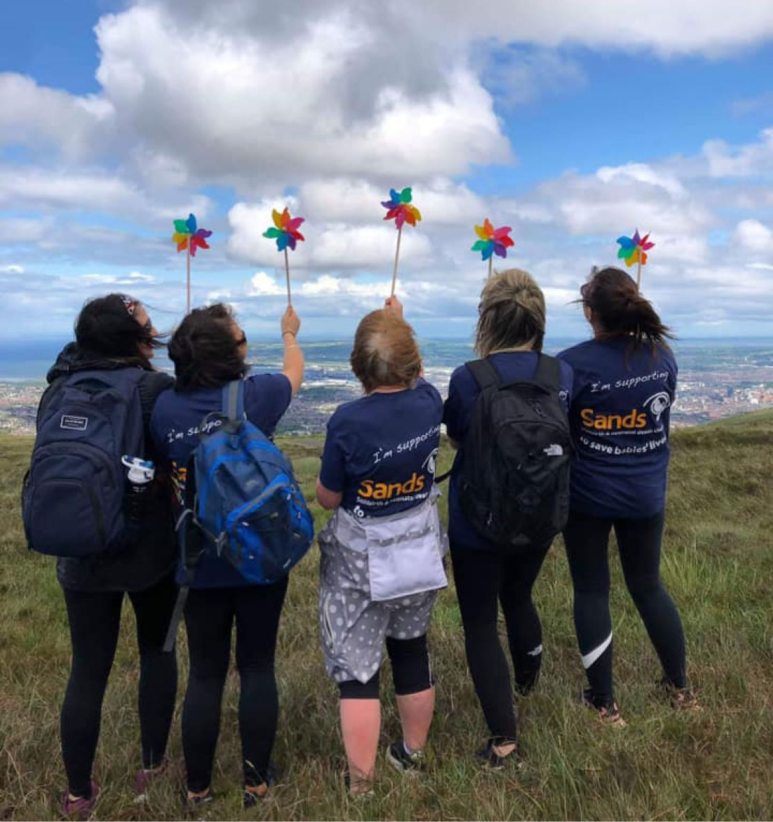 Five women wear Sands t-shirts while holding colourful wind spinners out to a grassy view.