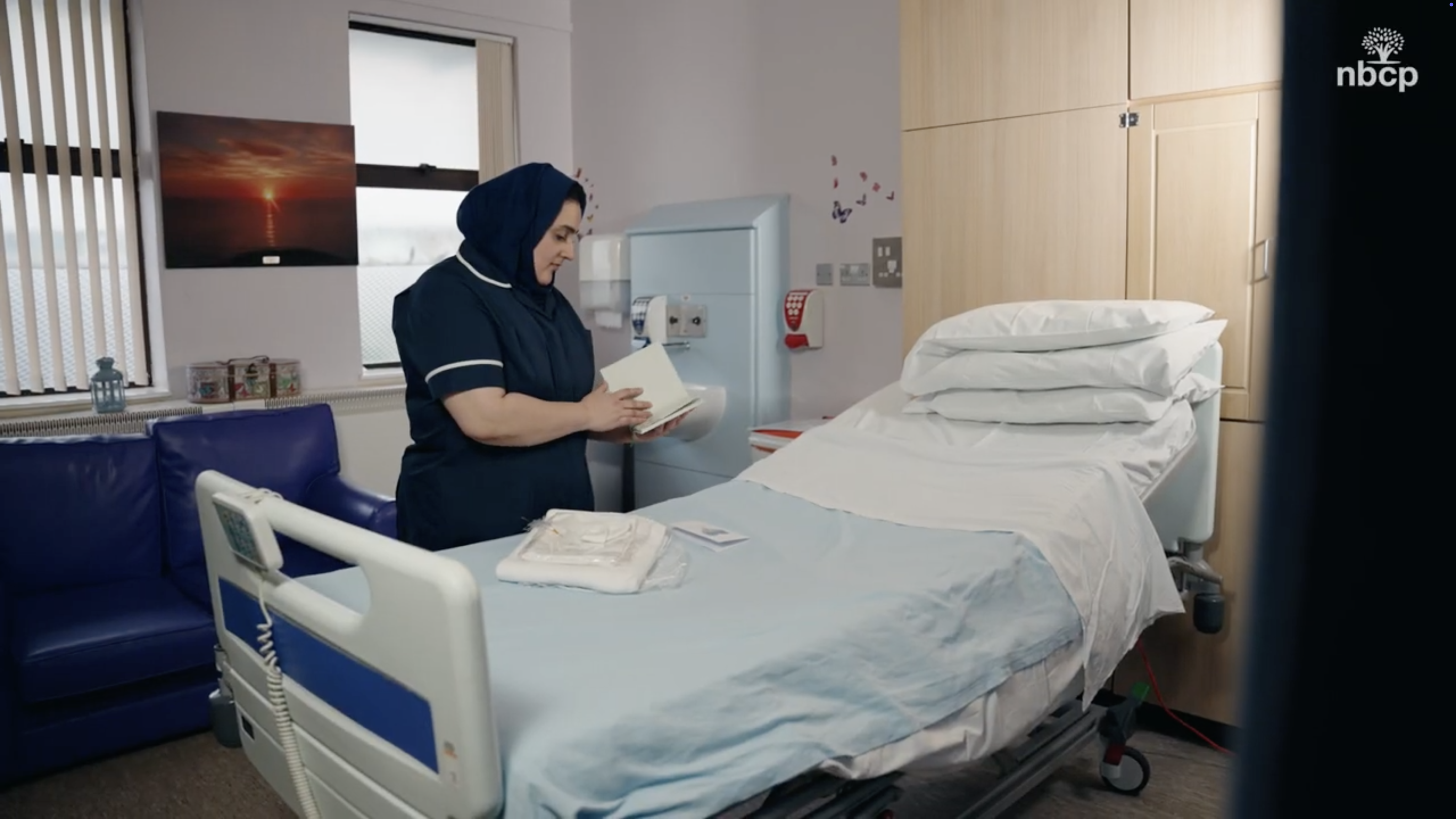 A nurse standing next to an empty hospital bed reading a booklet. She is wearing a dark navy blue nurse uniform and a headscarf.