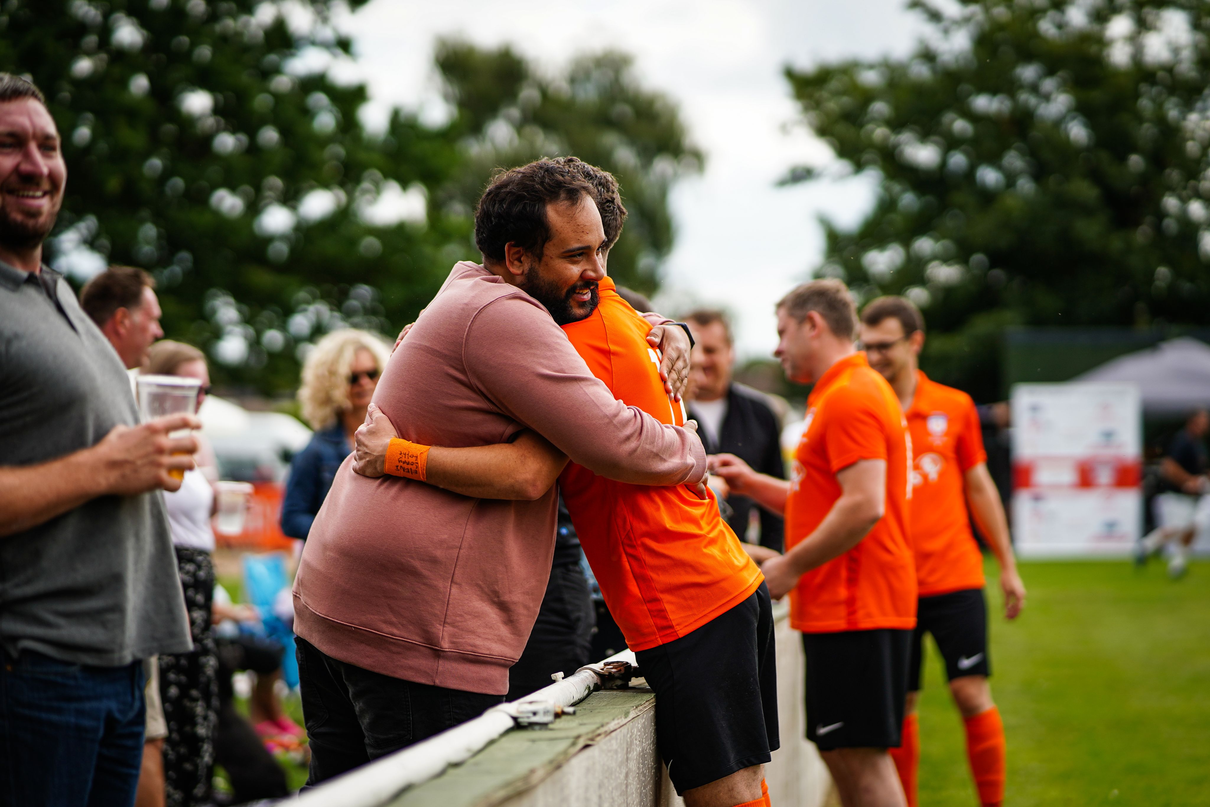 2 men hugging on the sidelines during a Sands United football match. One of the men is wearing a Sands United Kit while the other is in normal clothes.