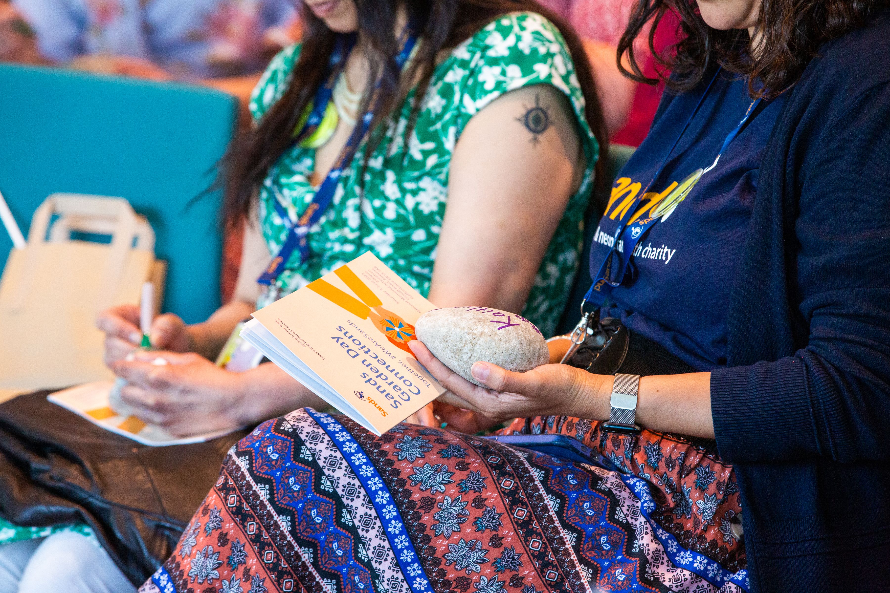 Two bereaved mothers holding stones with their babies name on them. The mother on the left is writing on the stone. The mother on the right is holding the stone and the Sands Garden Day Order of Service.