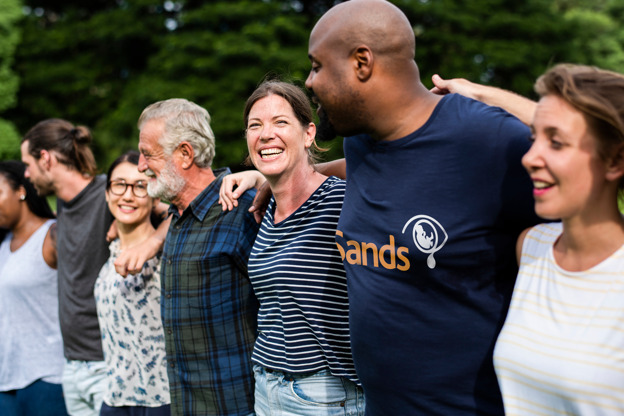 A group of people, one in a Sands top, stand smiling arm in arm.